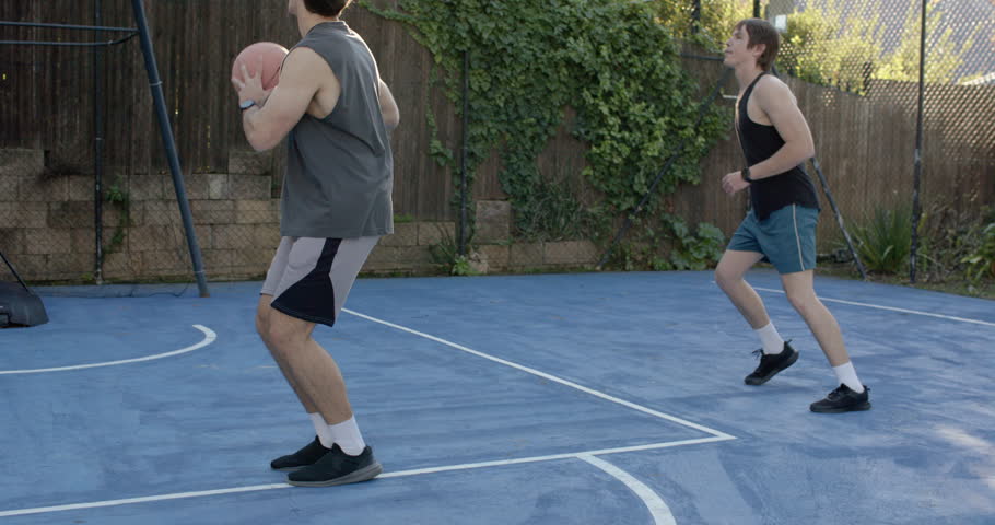 Three men on home portable-hoop court, gray shooter releasing ball, left man rebounding for score. Backyard, outdoor, basketball, lensflare, sunlight, backboard, net