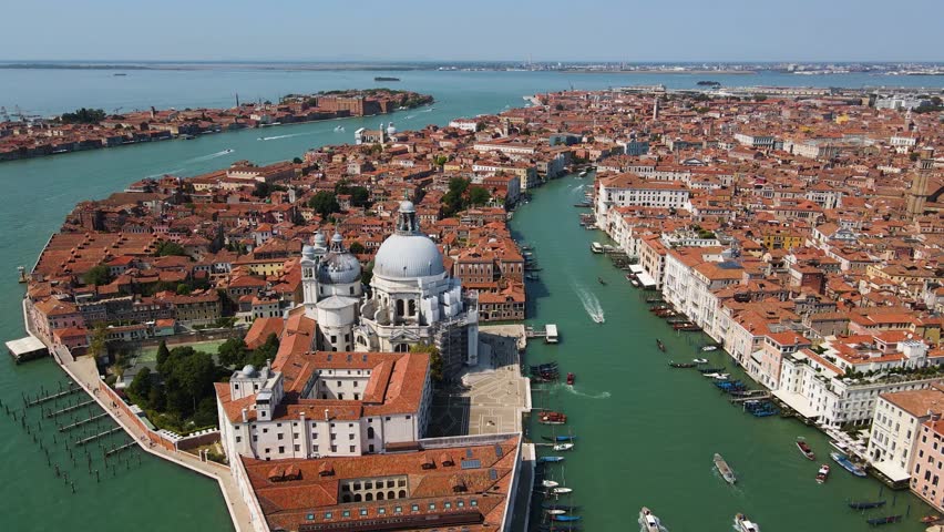 High altitude cinematic drone footage captures Grand Canal in Venice Italy showcasing iconic domes of Basilica di Santa Maria della Salute amidst vibrant terracotta rooftops and busy boat waterways.