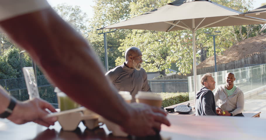 Diverse men watching mature man walking to counter after spotting takeaway cups, reading smartphone. Outdoor, cafe, bearded, friends, drinks, umbrella, sunny