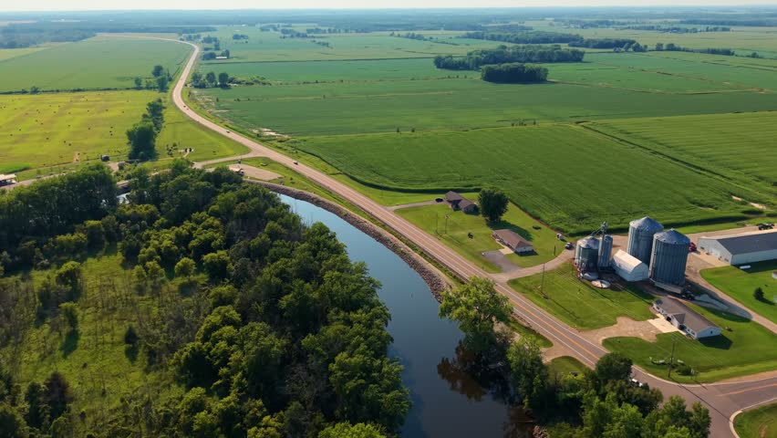 A high angle aerial shot capturing the Wolf river winding through lush green agricultural fields, farm silos, and country roads during a bright summer day in Shiocton, Wisconsin