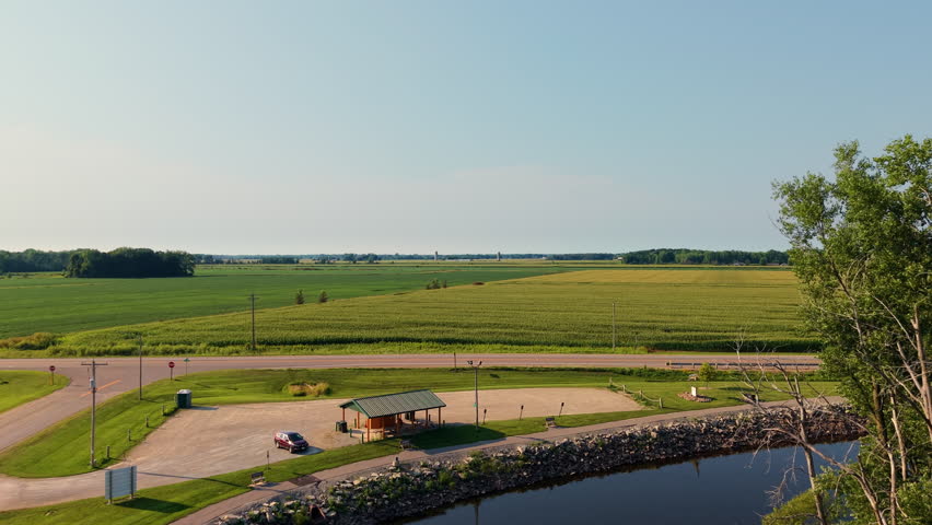 An aerial perspective of a peaceful countryside landscape featuring a winding river, agricultural fields, and industrial grain storage silos under a clear blue sky.