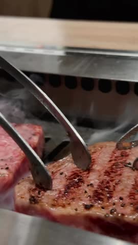 Close up shot of thick marbled beef steaks being grilled over an open flame on a hot grill, featuring rising smoke and metal tongs flipping the meat to achieve perfect sear marks.