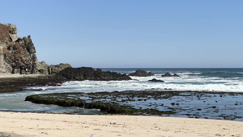 Waves crashing onto a rocky coast and sandy beach under a clear blue sky.