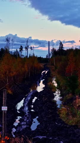 Aerial drone footage capturing wind turbine construction in rural landscape surrounded by colourful autumn forest and muddy road during twilight. Cinematic motion shot in remote wilderness.