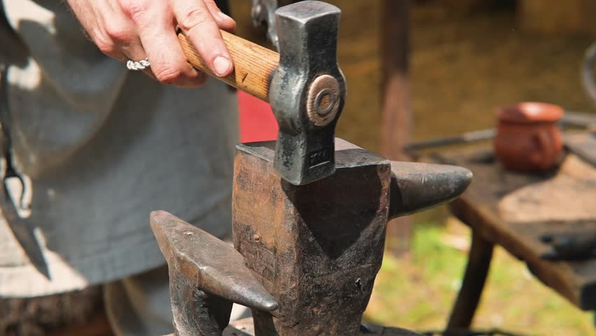 A man is hammering metal on an anvil in a reenactment setting. He is wearing simple clothing and focused on shaping the metal piece while tools are visible around him.