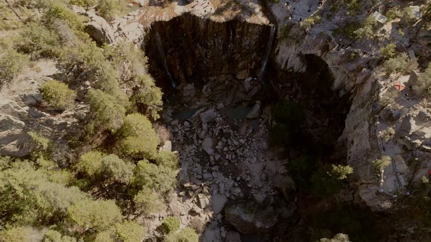 Aerial view of a rocky canyon featuring a seasonal waterfall, rugged cliffs, scattered boulders and dense forest, revealing dramatic natural terrain from above