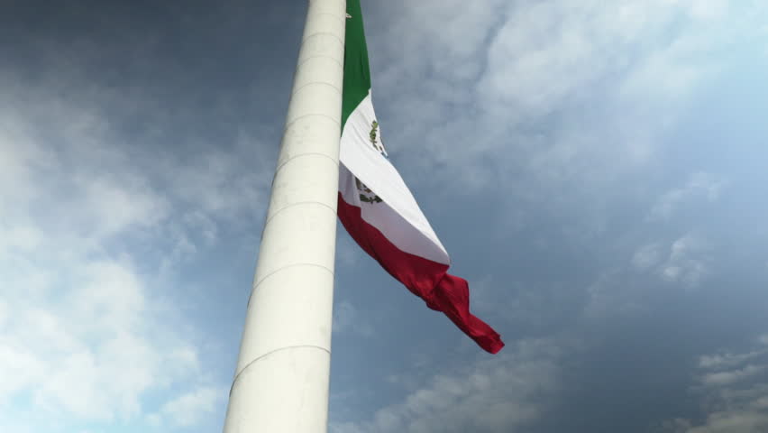 Cinematic low-angle shot looking up a massive white flagpole at the national tricolor flag of Mexico drooping down calmly against a dramatic backdrop of dark, overcast storm clouds.