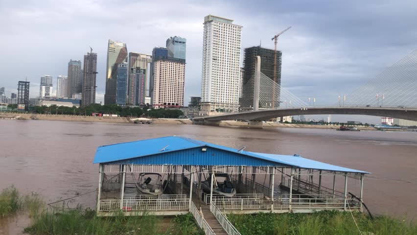 Panning view of riverside: blue-roofed boat dock, muddy river, modern high-rise skyline, and a cable-stayed bridge.