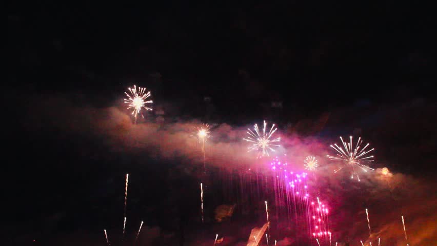 A large-scale public fireworks display at night, featuring low-lying white and pink bursts with accented green lighting at the bottom right.