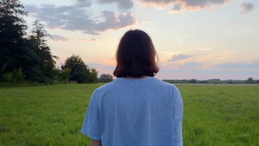 A young man stands in a grassy field. The young man looks towards the distant horizon and the colorful sunset in the background.