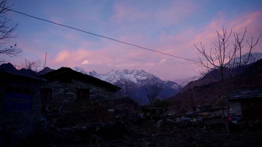 The silhouette of a stone farmhouse with a wooden ladder stands beneath bare branches, set against a pink and purple sunset over snow covered mountains in Nepal.