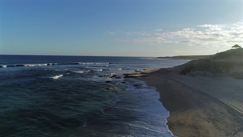 Drone flies low over the waterline at Playa El Rinconcito, passing rocks as ocean waves roll onto them, turning around a sand dune at the edge of the ocean toward the late afternoon light.