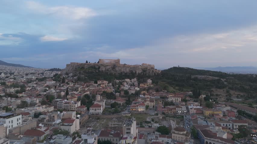 Acropolis of Athens crowns a rocky hill above dense city streets as a drone glides forward at dusk.