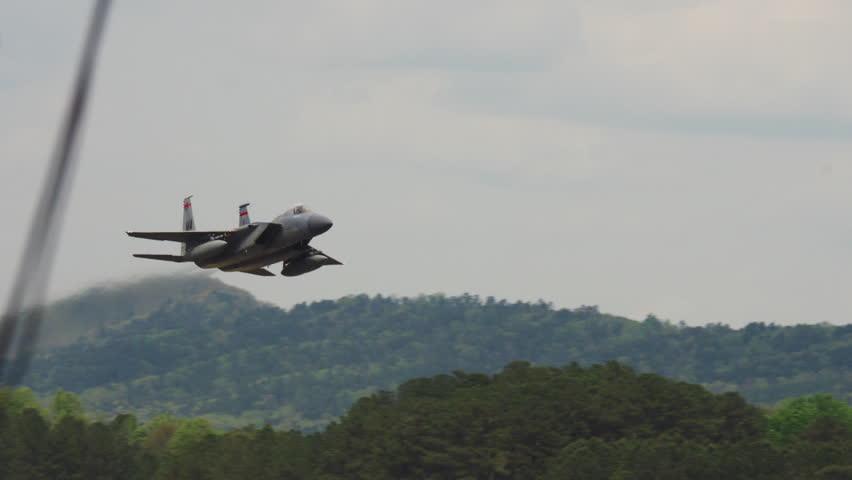 An F-15 fighter jet flies low over forested hills and into an overcast sky.