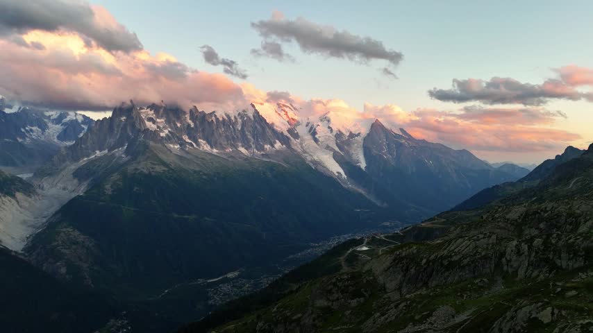 A wide view looks across the Chamonix valley in France toward jagged Mont Blanc peaks and glaciers, while pink sunset light colors snow, clouds and rocky slopes above the dark town below.