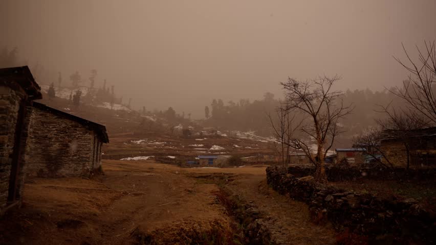A stone house stands in the foreground of a foggy mountain village, with scattered dwellings and snow patches on a terraced hillside behind it in Nepal.