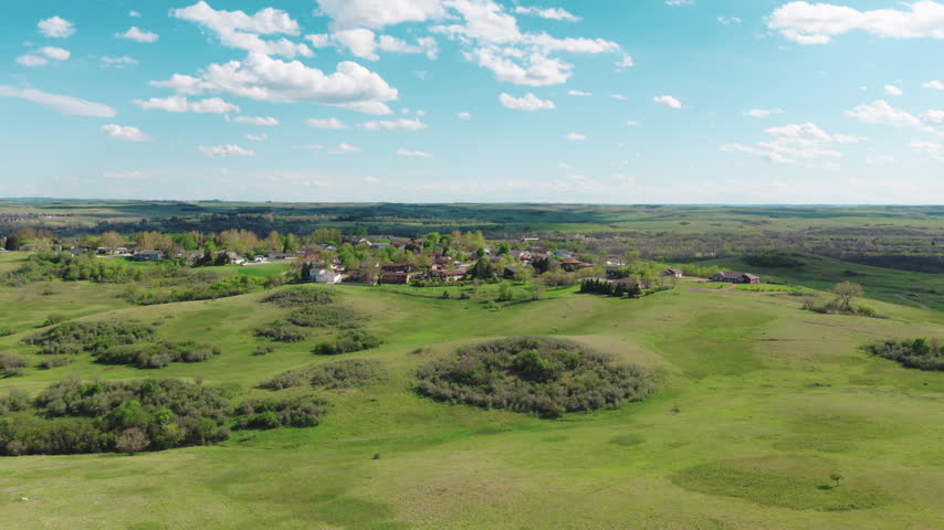 North Dakota showing a small town, rolling green prairie hills and scattered trees beneath a bright blue sky with puffy clouds.