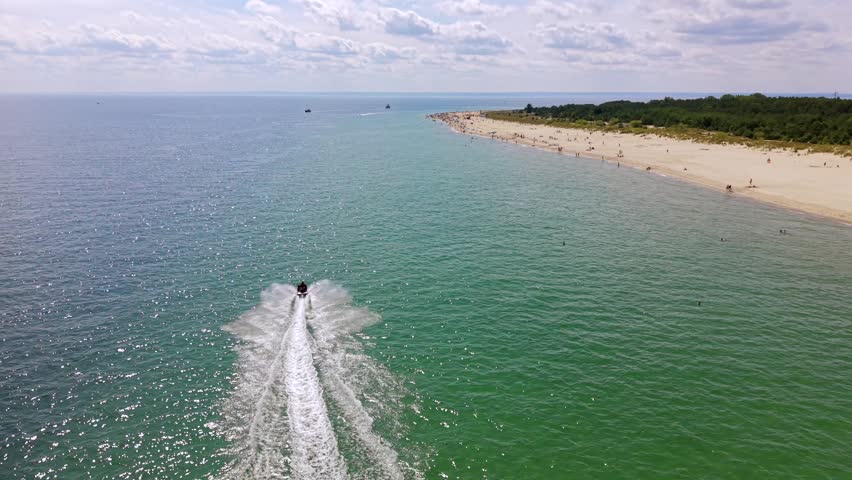 Aerial drone shot tracking a jet ski speeding across the turquoise Baltic Sea near the sandy beach in Hel on the Hel Peninsula in Poland, with a forested shoreline on a sunny summer day