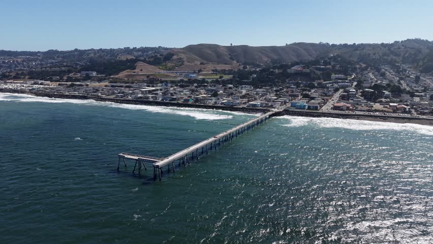 Expansive aerial view showing the Pacifica Municipal Pier extending into the Pacific Ocean, with the coastal city and rolling hills in the background on a bright, sunny day