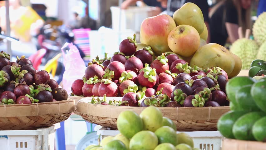 Vendor arranges fresh ripe mangosteen to a tropical fruit stall at an Asian market, with mango, avocado and durian in the background. Fresh produce shopping and local food market concept.