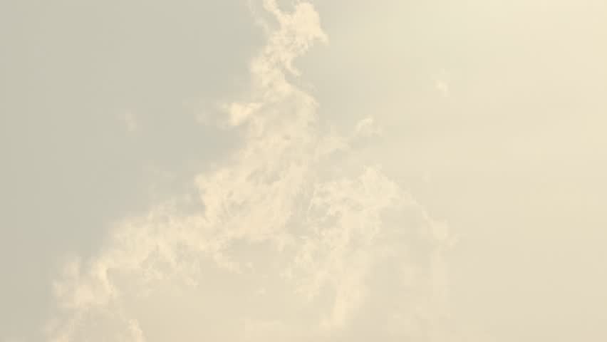 Wisps of white clouds drifting slowly against a pale, warm-toned sky in soft sunlight