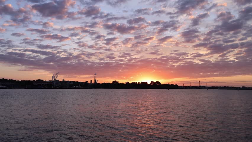 Time lapse of a spectacular summer sunrise over the IJ waterfront and port area in Amsterdam, Netherlands. Colorful morning sky with dramatic clouds reflecting warm sunlight over the water.