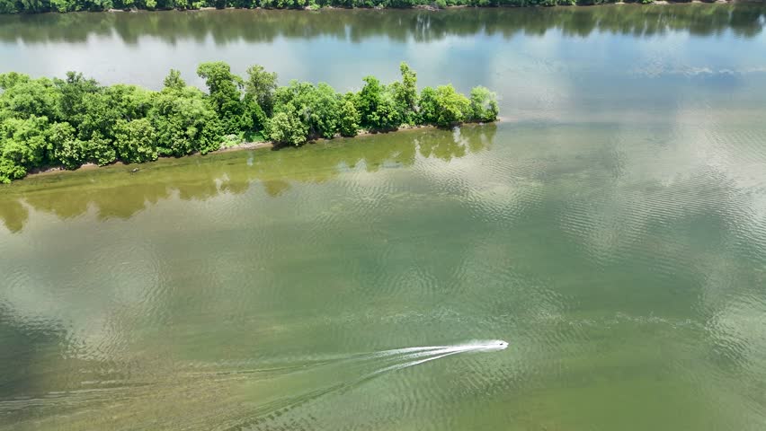 High-angle aerial drone video of a personal watercraft cruising across the calm Potomac river at Algonkian Regional Park in Sterling, VA. The small boat creates a prominent white wake as it navigates past a lush, green wooded island. Sunlit sky and soft cloud reflections pattern the water surface. Ideal stock footage for summer outdoor recreation, water sports, river activities, travel marketing, regional tourism, and leisure lifestyle storytelling. Loudoun County, Virginia.