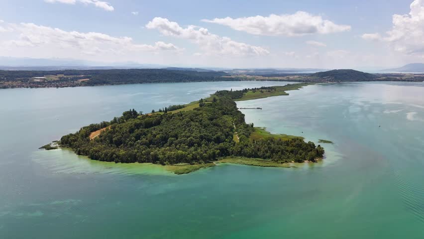 An aerial drone overlooks St Peters Island in Lake Biel, Switzerland, showing its wooded shoreline, shallow turquoise water, narrow peninsula, distant hills, and scattered clouds.