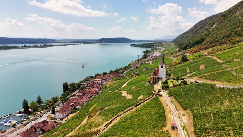 An aerial drone moves along the vineyard slopes of Ligerz beside Lake Biel, Switzerland, passing a hillside church, lakeside homes, terraced vines, and a boat cutting across blue water.
