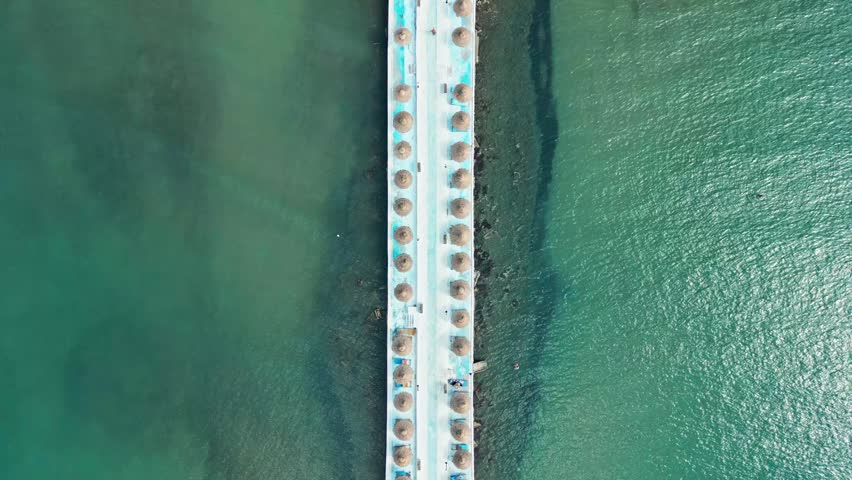 Albania, Durres 07 09 2025: Top-down vertical drone video of a minimalist pathway over the sea connecting to Hawaii Beach Club, lined on both sides with straw umbrellas.
