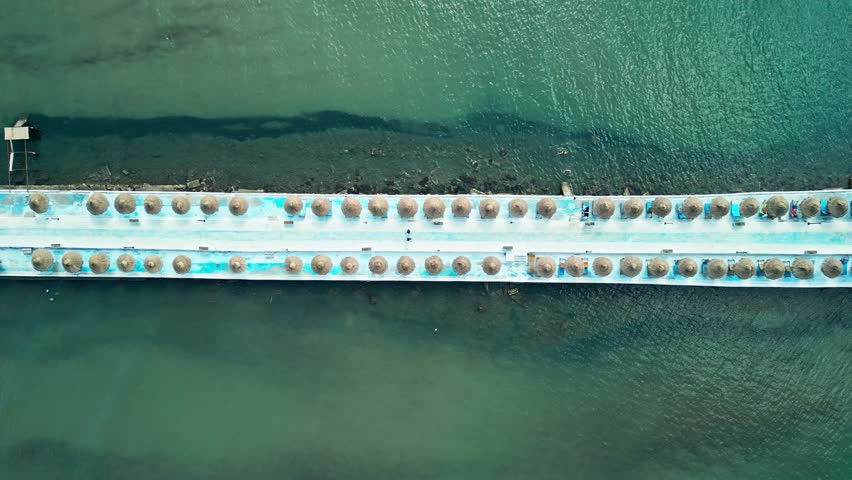 Albania, Durres 07 09 2025: Top-down drone video of a horizontal pedestrian pathway over the sea connecting Durres beach to Hawaii Beach Club, lined with straw umbrellas.