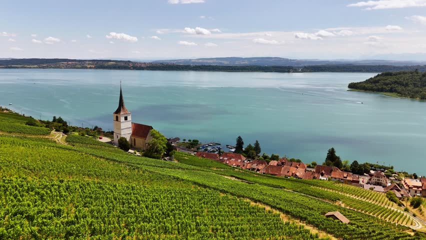 An aerial drone approaches the hillside church in Ligerz, Switzerland, moving above terraced vineyards and red-roofed homes with Lake Biel and distant wooded shores filling the background.