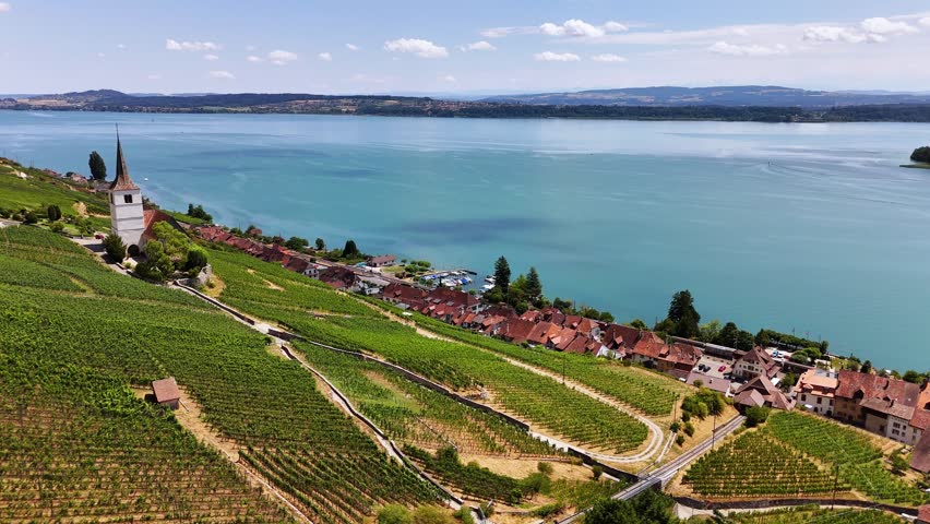 An aerial drone pulls back from Ligerz church across terraced vineyards, revealing more red-roofed homes and hillside paths beside the broad turquoise water of Lake Biel in Switzerland.