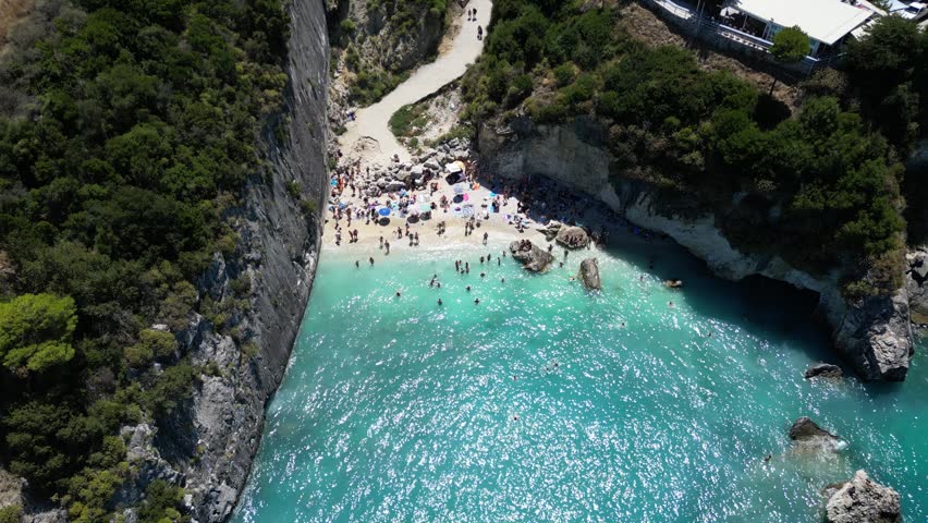 Aerial view of Porto Limnionas beach with turquoise water, rocky cliffs, and beachgoers under umbrellas in Zakinthos, Greece.