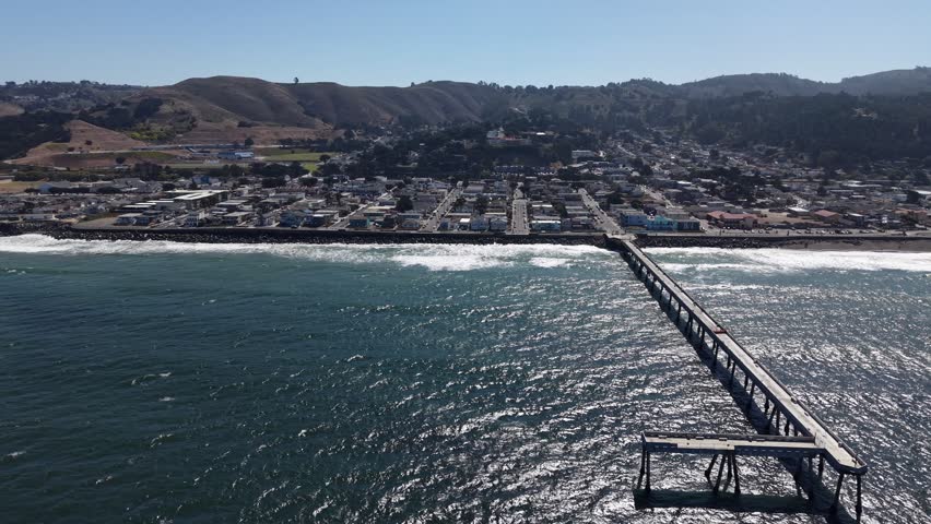 Aerial perspective of the historic Pacifica Municipal Pier stretching into the glistening Pacific Ocean, with the coastal cityscape and rolling hills of Northern California in the background