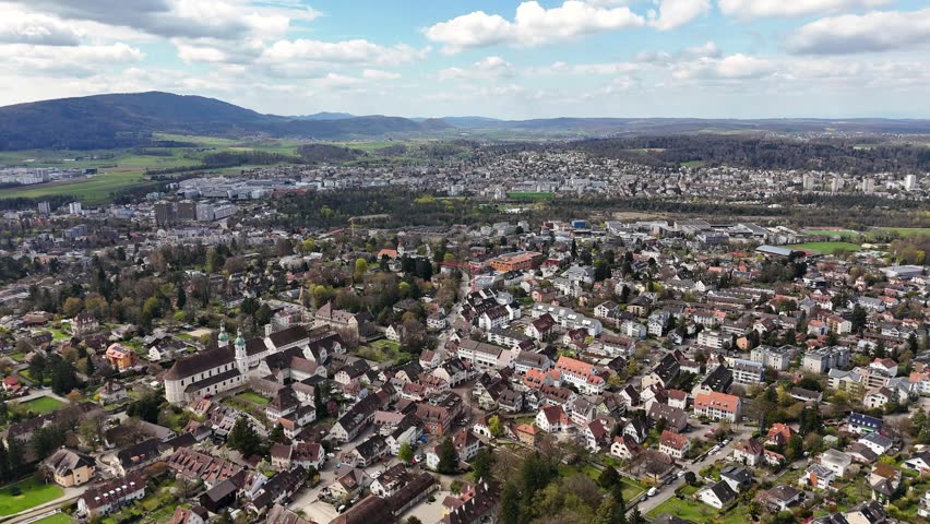 Aerial drone glides over Arlesheim in Switzerland, showing a prominent church, tiled rooftops, residential streets, green fields, and wooded hills beneath scattered clouds.