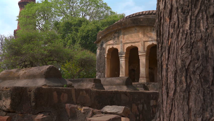 Tracking shot of an ancient arched stone pavilion and historic tombs framed by a tree trunk at Qutub complex in New Delhi, India.