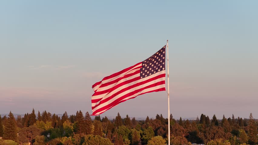 American Flag Waving On Flagpole Above Tree Canopy At Sunset