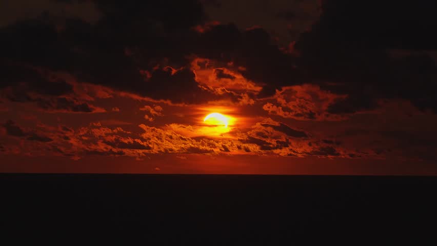 A partial solar eclipse occurs during sunset over the dark ocean. The glowing sun is partially covered by the moon amidst dark clouds.