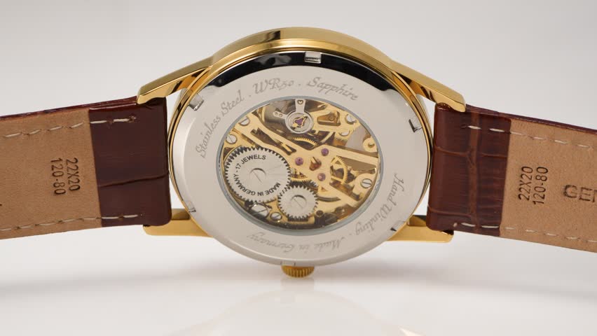 Close-up footage of a luxury mechanical wristwatch featuring a transparent exhibition caseback, visible gears and movement, gold tone case and brown leather strap on a clean light background.