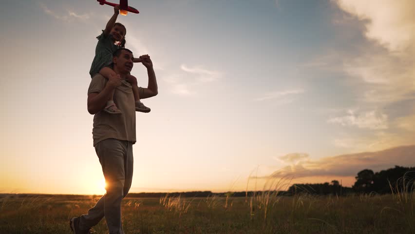 Father carries child on shoulders through grassy field at sunset. Happy child holds toy plane high in air. Parent walks across meadow as family enjoys outdoor play. Sunset glows behind them.