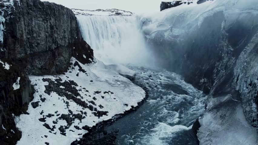 Dramatic aerial drone view of the massive Dettifoss waterfall rushing through a rugged, snow-dusted rock canyon with icy mist rising in winter.