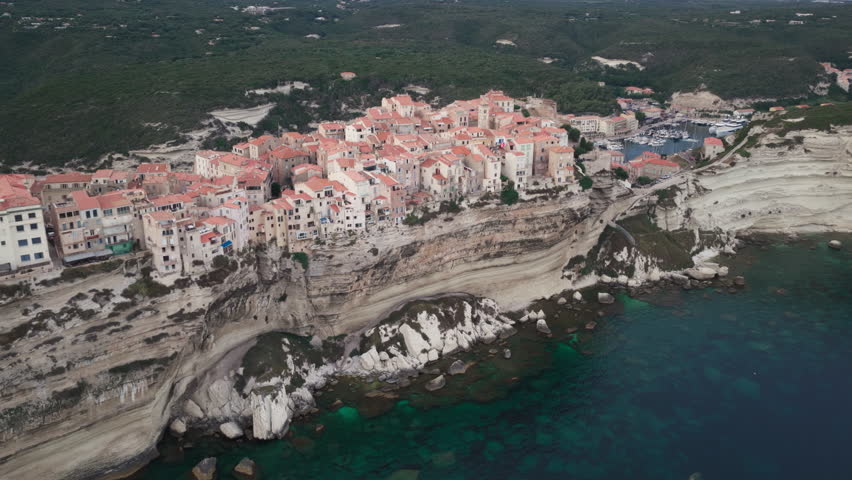 A sunset aerial over epic limestone cliffs and historic buildings of old town Bonifacio in Corsica, France with luxury yachts in the protected harbor and the turquoise blue water of the Mediterranean.