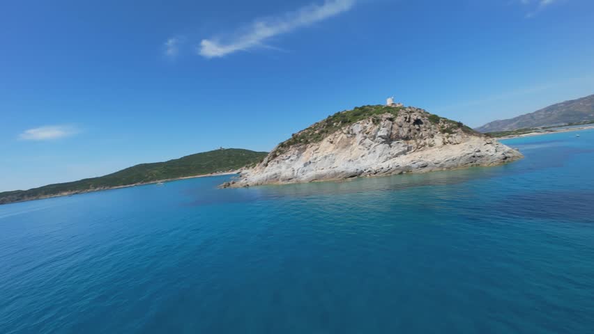 An FPV drone flying low along rocky cliffs above turquoise water, anchored boats and sandy beach in the distance, Villasimius, Sardinia. FPV aerial footage of Mediterranean coast.
