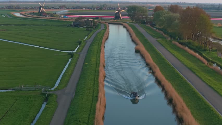 Majestic aerial drone shot following a boat cruising along a serene canal in the netherlands, with traditional windmills in the background