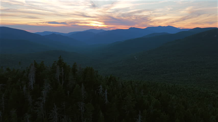 Slow aerial pan above evergreen treetops reveals a vast forested valley and layered White Mountain ridges beneath a dramatic summer sunset along New Hampshire’s Kancamagus Highway. Text or logo room.