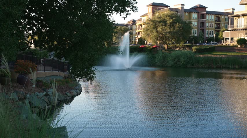Large fountain sprays across urban pond beside residential apartment buildings, with trees, landscaped greenery and warm reflections creating a peaceful waterfront scene