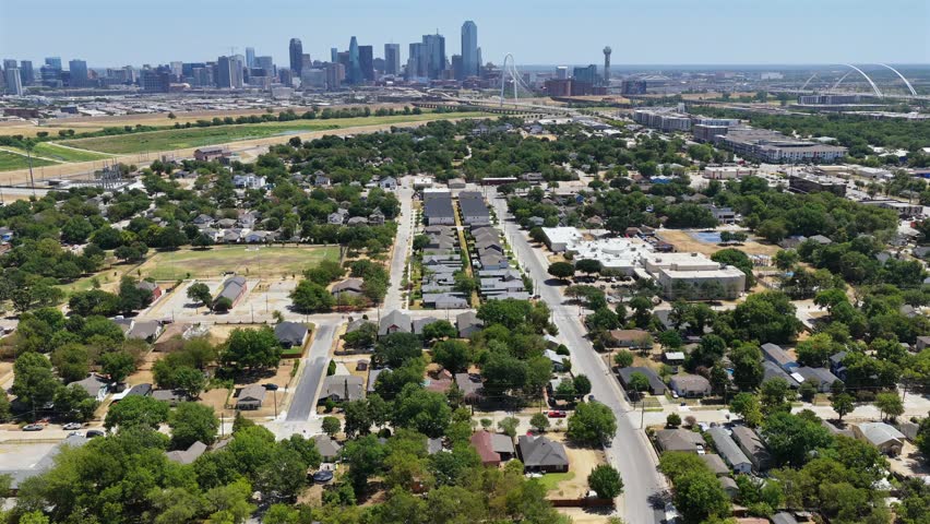 A high summer aerial looks across West Dallas bungalows, vacant parcels, new infill construction, and a long residential street toward Downtown Dallas. Reunion Tower and the paired white arches of the Margaret McDermott Bridge mark the horizon across the Trinity River floodway.