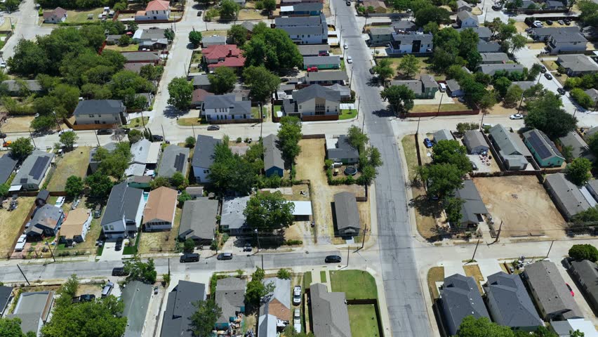 The drone rises and tilts from a West Dallas residential intersection to follow a long neighborhood street toward Downtown. Older bungalows, new infill homes, vacant parcels, apartment rows, and mature trees lead to the skyline, Reunion Tower, and the Margaret McDermott Bridge on the horizon.