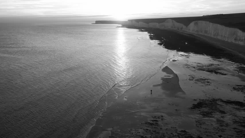 Aerial drone view showing chalk cliffs, coastline and calm sea with sunlight reflecting across water beneath cloudy sky in Brighton, England, United Kingdom
